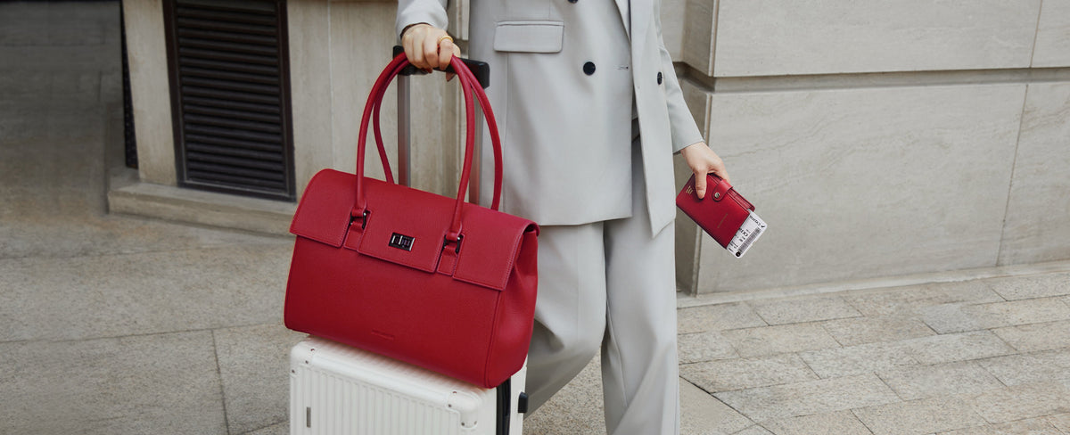 Model moving through an airport with the Victoria Hyde Amal garnet red convertible business bag for 16-inch laptop secured to the handle of a white suitcase using the integrated luggage strap