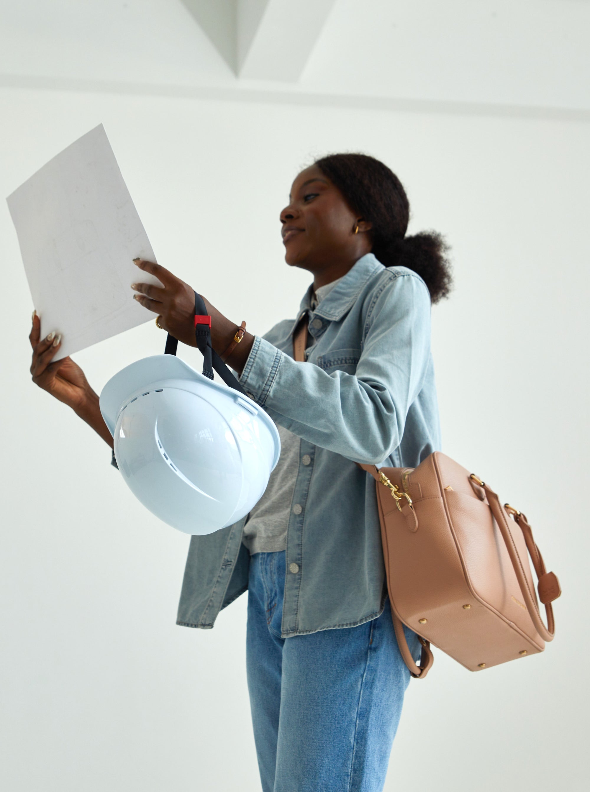 Frau mit lockigem Haar hält weiße Schutzhelm und braune Victoria Hyde Handtasche, prüft Baupläne in minimalistischem Raum – moderne, nachhaltige Damenhandtasche für Business, Architektur oder Bauwesen.