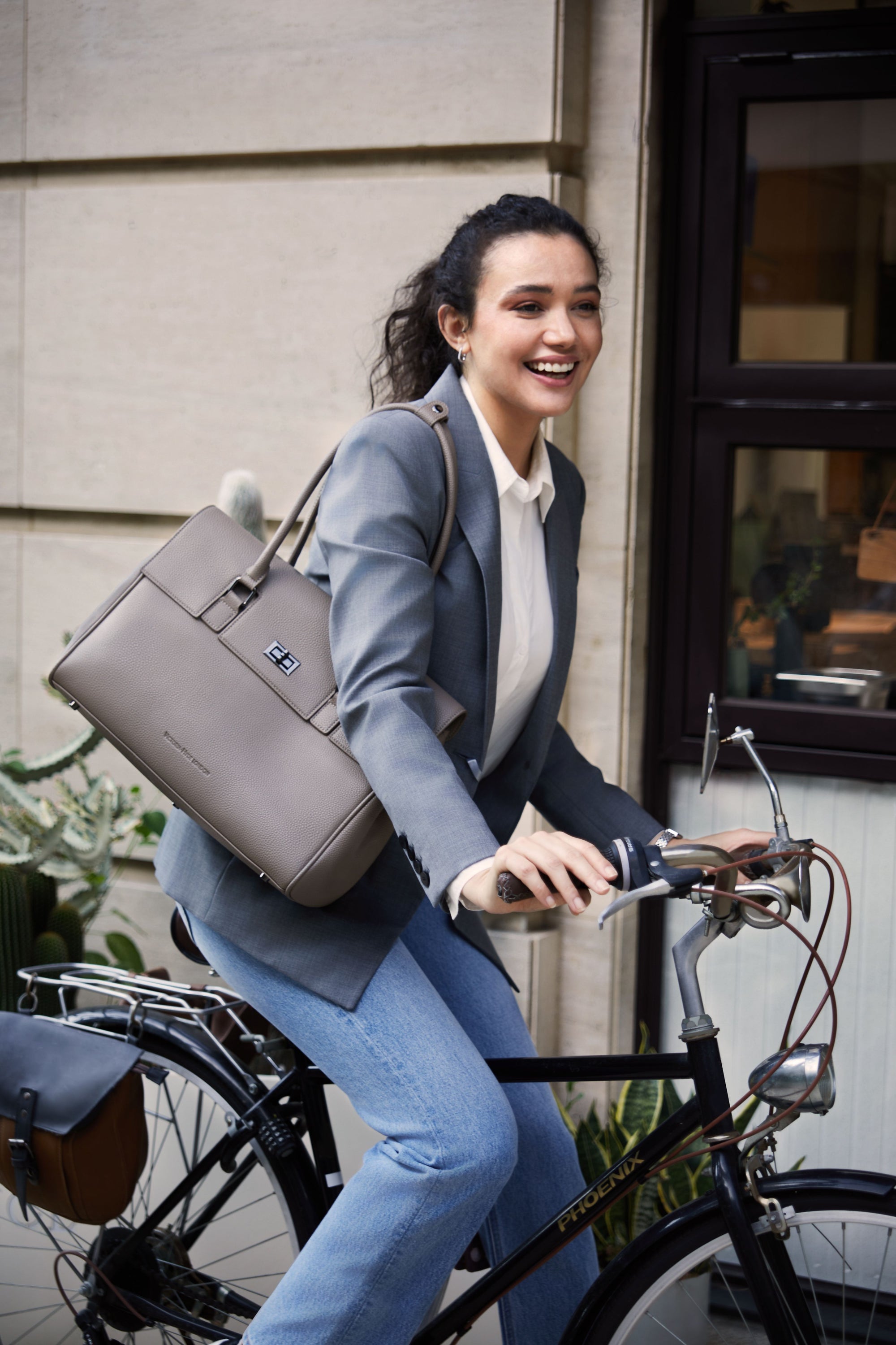Frau mit lockigem Haar fährt Fahrrad in urbaner Umgebung, trägt grauen Blazer und große graue Victoria Hyde Handtasche – stilvolle, nachhaltige Damenhandtasche für Business und Freizeit.