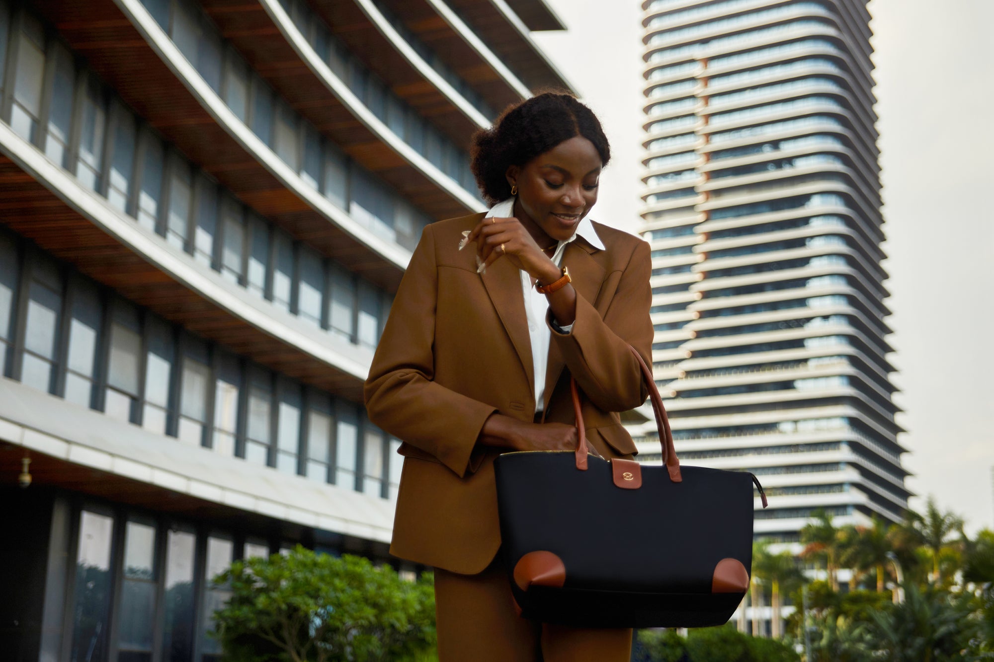 Frau im braunen Anzug mit weißem Hemd und großer schwarzer Victoria Hyde Handtasche mit braunen Akzenten, lächelnd in urbaner Umgebung mit moderner Architektur – stilvolle, nachhaltige Damenhandtasche für Business und Alltag.
