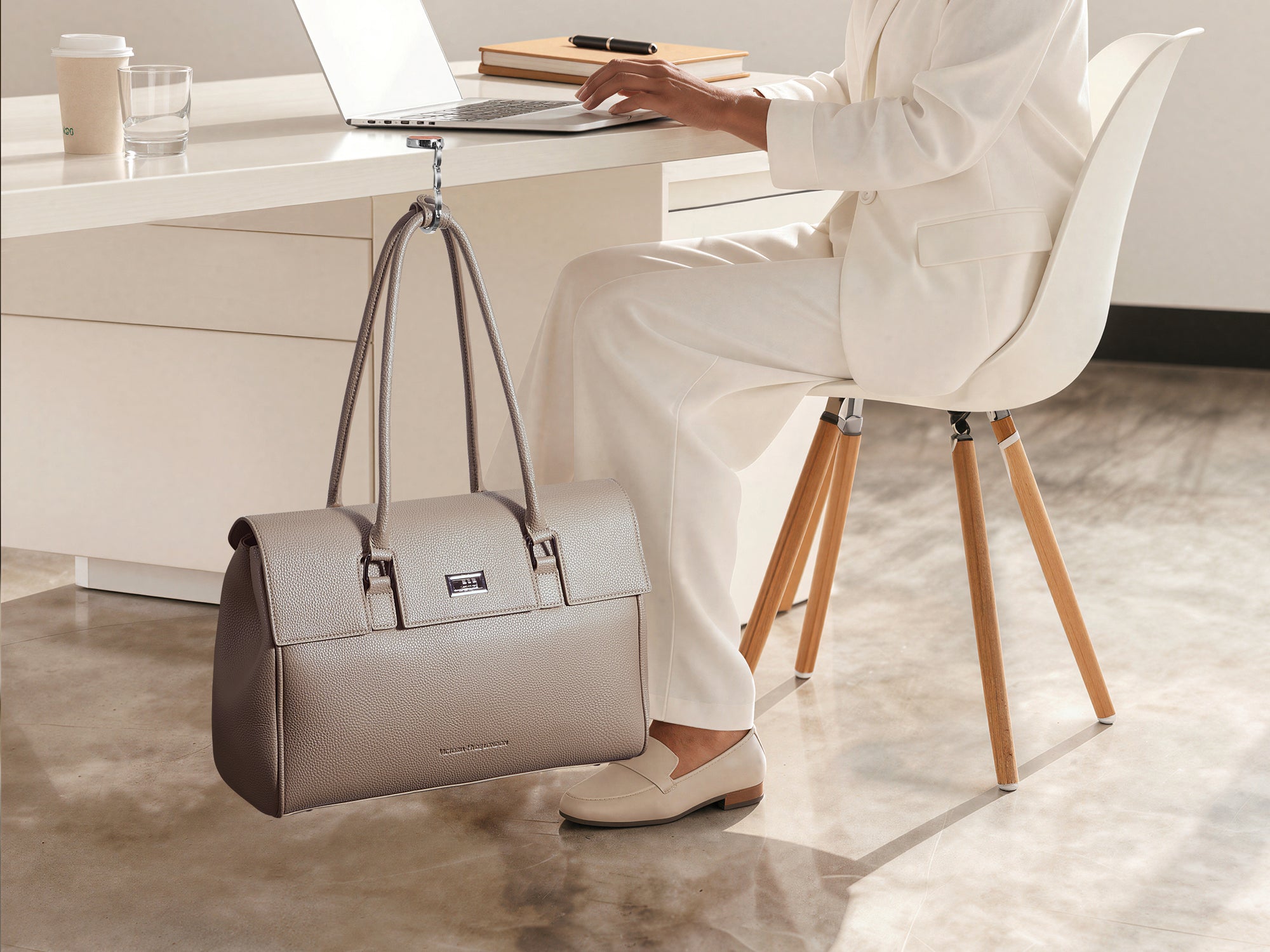 Eine Frau im weißen Hosenanzug arbeitet am Schreibtisch im Büro, während ihre taupefarbene Victoria Hyde Jolene Laptoptasche mit einem Handtaschenhalter ordentlich an der Tischkante hängt.