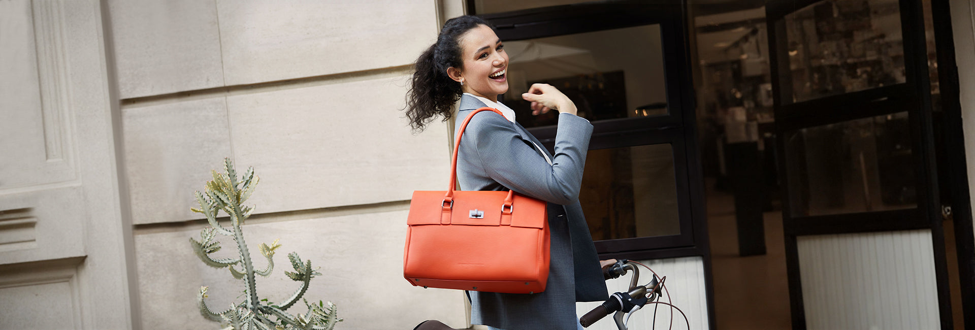 Eine lächelnde Frau im grauen Business-Blazer fährt Fahrrad in der Stadt mit der eleganten Victoria Hyde Jolene Laptoptasche in Orange.