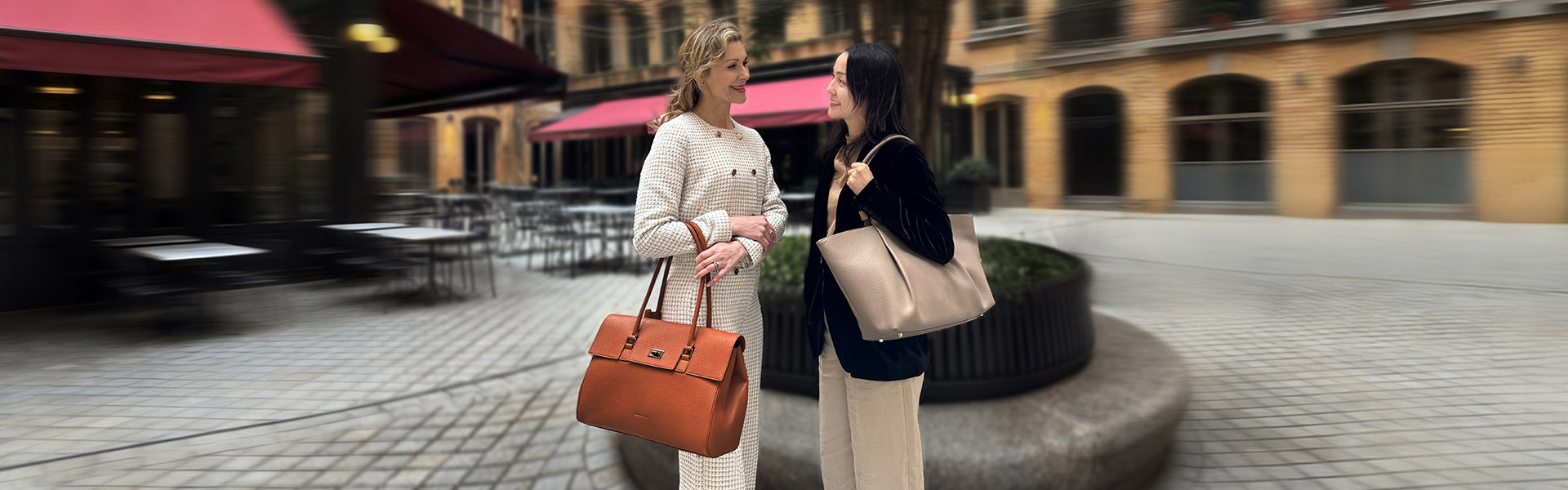 Zwei stilvolle Frauen unterhalten sich auf einem europäischen Stadtplatz beim Pendeln mit der orangefarbenen Jolene Laptoptasche und einer beigen Victoria Hyde Work Tote Bag.