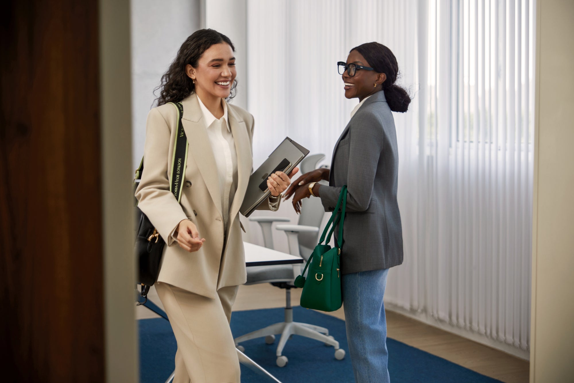 Zwei lächelnde Business-Frauen im eleganten Hosenanzug und Blazer unterhalten sich im modernen Büro mit ihren Victoria Hyde New Work Laptoptaschen für Damen.
